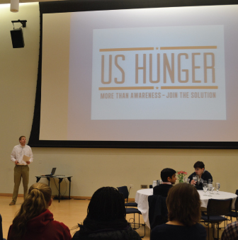 Students sit at tables, while a presenter discusses hunger in the US